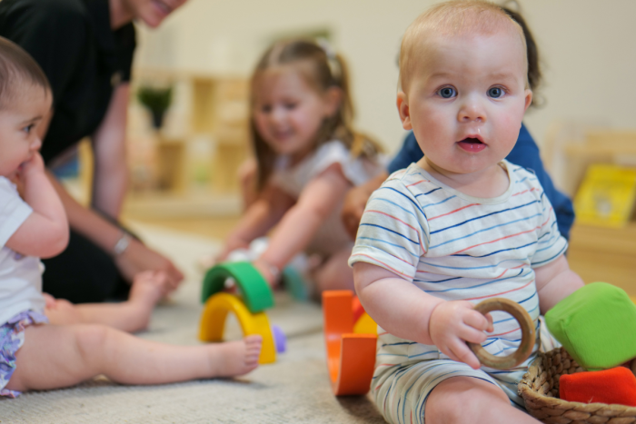 Baby in childcare centre holding round rattle with educator and children in background